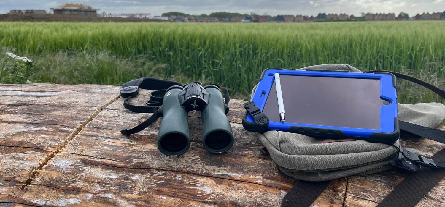 Ecologist binoculars and a tablet with pen on a green rucksack lying on a wooden surface with a meadow and in the bakcground